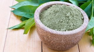 A bowl of henna powder with fresh henna leaves on a wooden table